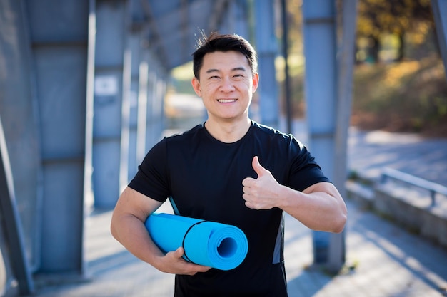 Portrait d'athlète asiatique avec tapis pour lui près du stade regardant la caméra et souriant tenant le pouce levé instructeur de yoga