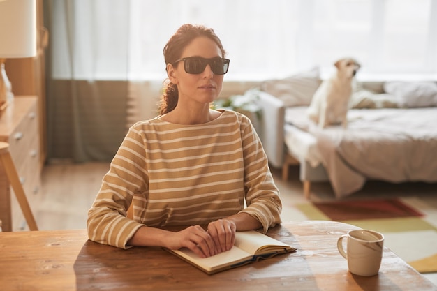 Portrait aux tons chauds d'une femme aveugle moderne lisant un livre en braille assis à table dans un intérieur confortable avec un chien-guide en arrière-plan, espace pour copie