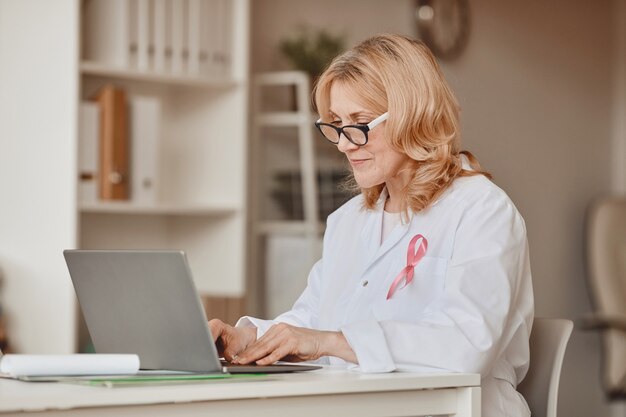 Portrait aux tons chauds de femme médecin mature avec ruban rose a effectué le tombé sur une blouse blanche tout en travaillant à un ordinateur portable dans un bureau moderne, symbole de sensibilisation au cancer du sein, espace de copie