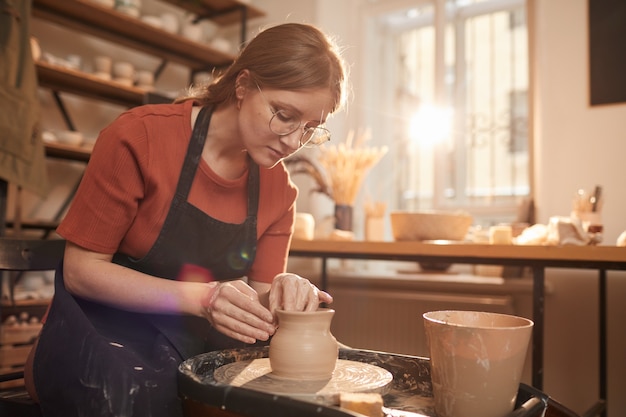 Portrait aux tons chauds d'une jeune artisane façonnant de l'argile sur un tour de poterie dans un atelier ensoleillé et profitant des arts et de l'artisanat, espace de copie