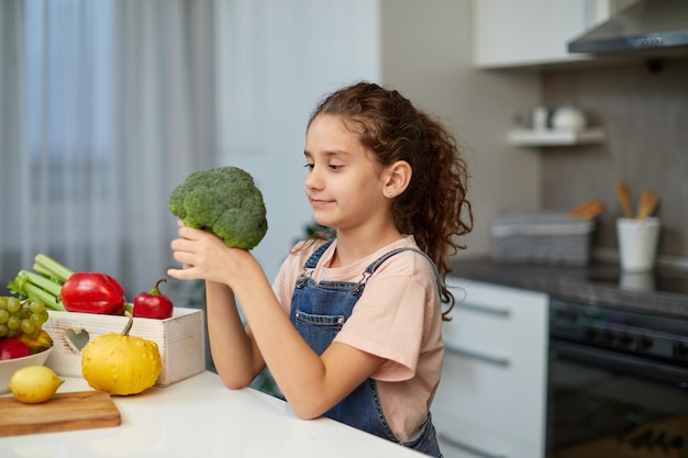 Portrait avant d'une petite fille aux cheveux bouclés, vêtue d'un jean et d'un t-shirt, tenant et regardant le brocoli, assise à table dans la cuisine.