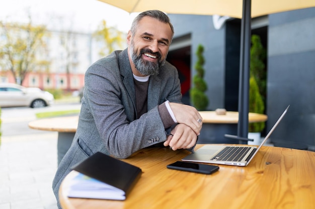 Portrait d'un avocat adulte mature aux cheveux gris et à la barbe assis sur une terrasse avec un ordinateur portable