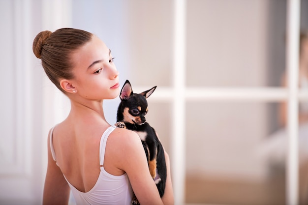 Portrait d'une ballerine en blanc avec un petit chien chihuahua dans ses mains dans une belle salle blanche devant un miroir.