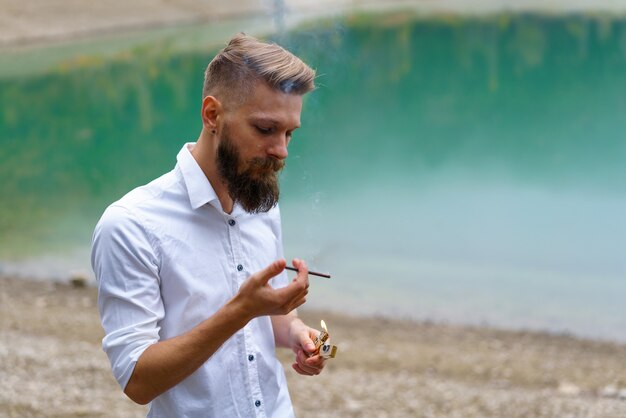 Portrait d'un beau barbu allume une cigarette avec un briquet à l'extérieur sur fond de...