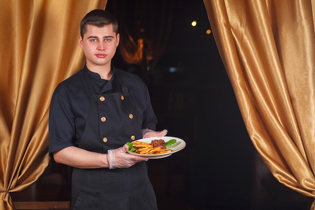 Portrait d'un beau chef cuisinier masculin souriant en uniforme debout avec un délicieux plat à la cuisine. Chef masculin tenant une assiette avec un plat cuisiné.
