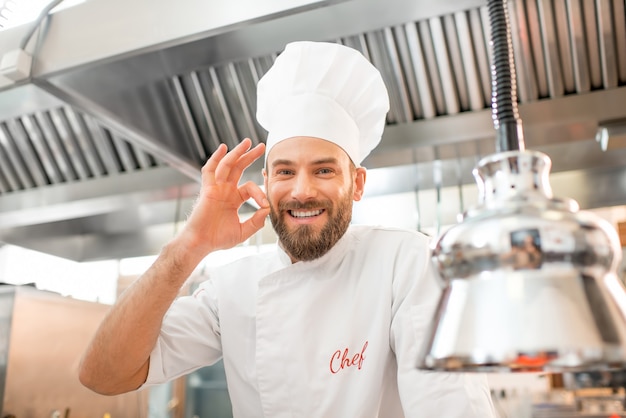 Portrait d'un beau chef cuisinier en uniforme montrant un délicieux signe dans la cuisine du restaurant