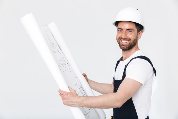 Portrait d'un beau constructeur barbu portant une salopette debout isolé sur un mur blanc, regardant le projet