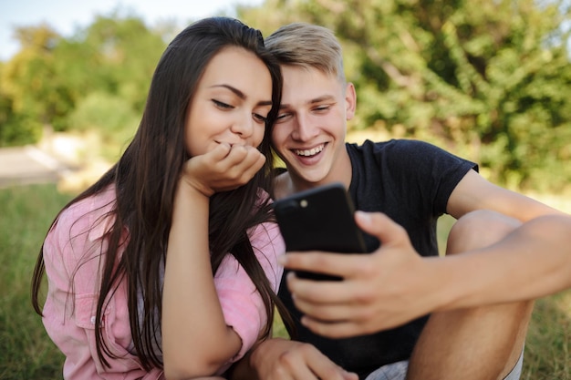 Portrait d'un beau couple souriant assis sur la pelouse dans le parc et utilisant joyeusement un téléphone portable ensemble