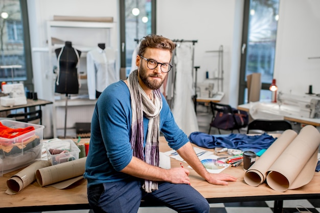 Portrait d'un beau créateur de mode assis avec des croquis en papier au studio plein d'outils et de vêtements de couture