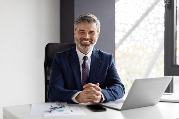 Portrait d'un beau homme d'affaires d'âge moyen souriant assis sur le lieu de travail au bureau