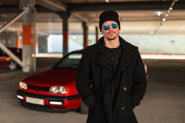 Portrait d'un beau jeune conducteur avec des lunettes de soleil dans un manteau à la mode avec un chapeau près d'une voiture rouge dans un parking