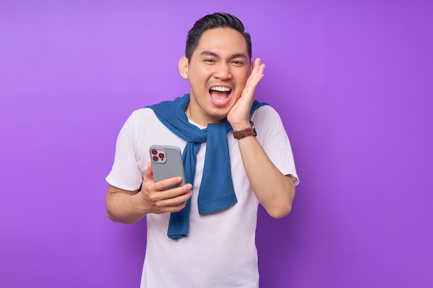 Portrait d'un beau jeune excité dans des vêtements décontractés tenant un smartphone isolé sur fond violet