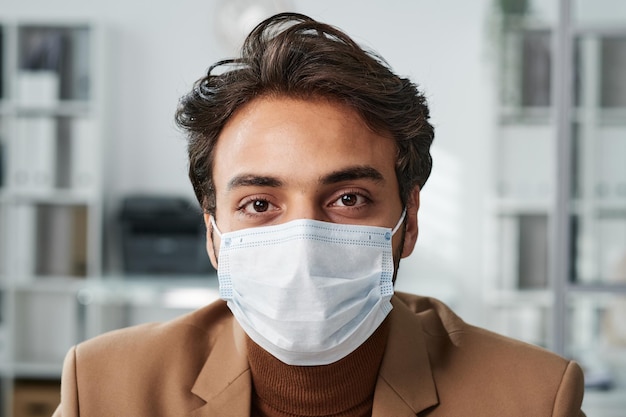 Portrait de beau jeune gestionnaire du Moyen-Orient en masque assis dans un bureau contemporain