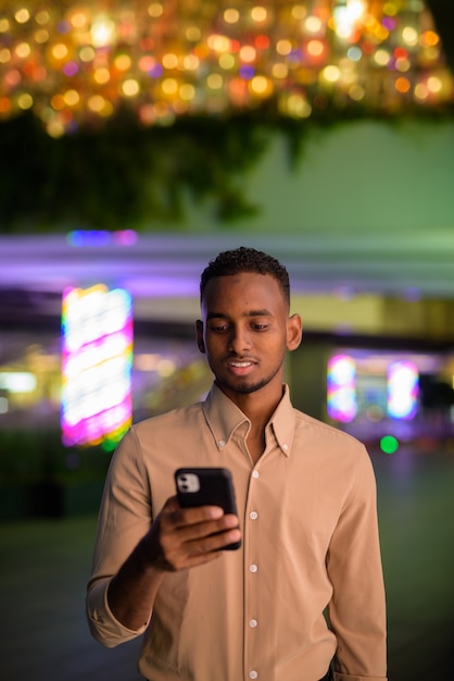 Portrait de beau jeune homme d'affaires africain noir portant des vêtements décontractés à l'extérieur en ville et à l'aide de téléphone mobile