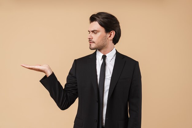 Portrait d'un beau jeune homme d'affaires confiant portant un costume debout isolé sur un mur beige, présentant un espace de copie