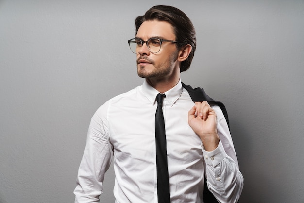 Portrait d'un beau jeune homme d'affaires confiant portant un costume debout isolé sur un mur gris, posant avec une veste sur son épaule