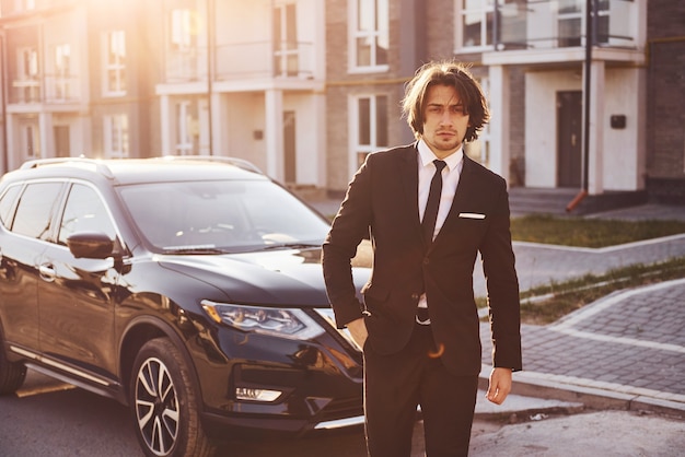 Portrait de beau jeune homme d'affaires en costume noir et cravate à l'extérieur près d'une voiture moderne dans la ville.