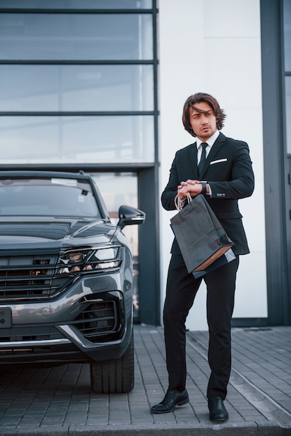 Portrait de beau jeune homme d'affaires en costume noir et cravate à l'extérieur près d'une voiture moderne et avec des sacs à provisions.