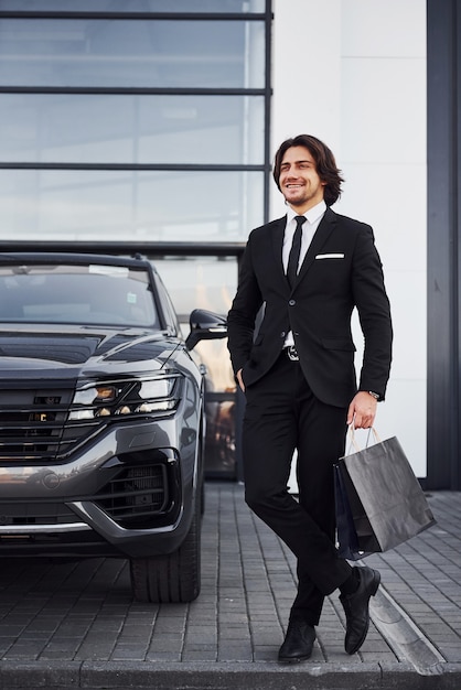 Portrait de beau jeune homme d'affaires en costume noir et cravate à l'extérieur près d'une voiture moderne et avec des sacs à provisions.
