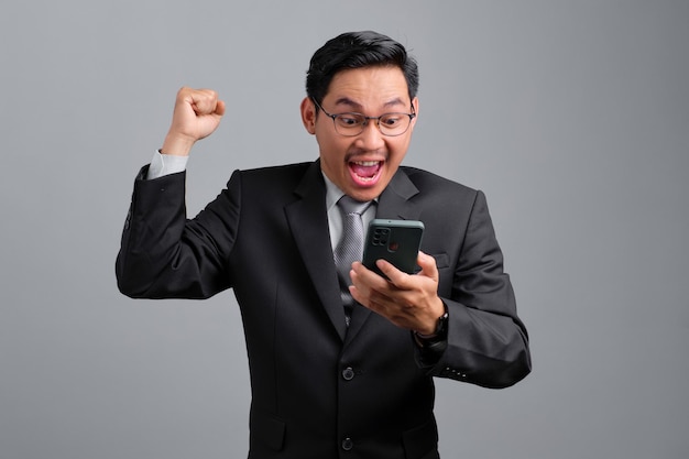 Portrait d'un beau jeune homme d'affaires excité en costume formel et lunettes montrant le geste du gagnant lisant de bonnes nouvelles sur un téléphone portable isolé sur fond gris