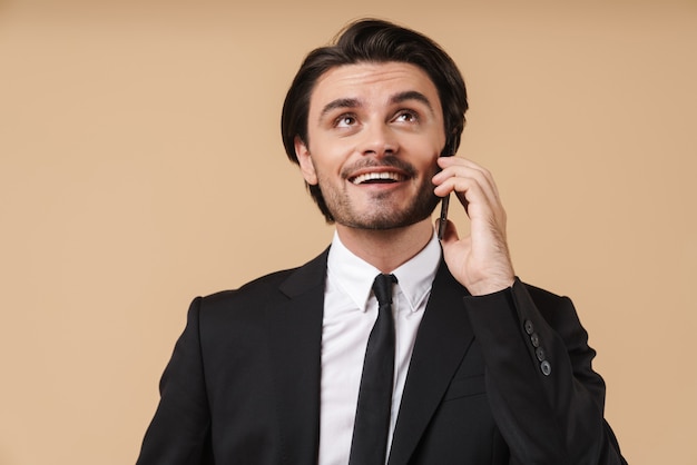 Portrait d'un beau jeune homme d'affaires souriant portant un costume debout isolé sur un mur beige, parlant au téléphone portable