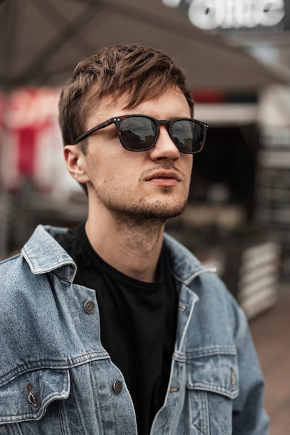 Portrait d'un beau jeune homme américain avec une barbe dans des lunettes de soleil à la mode dans une élégante veste en jean bleu dans un t-shirt dans la rue un jour de printemps. Beau mec hipster se promène dans la ville.