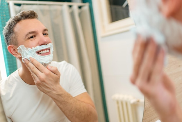 Portrait d'un beau jeune homme appliquant de la crème à raser devant le miroir de sa salle de bain. Mise au point sélective.