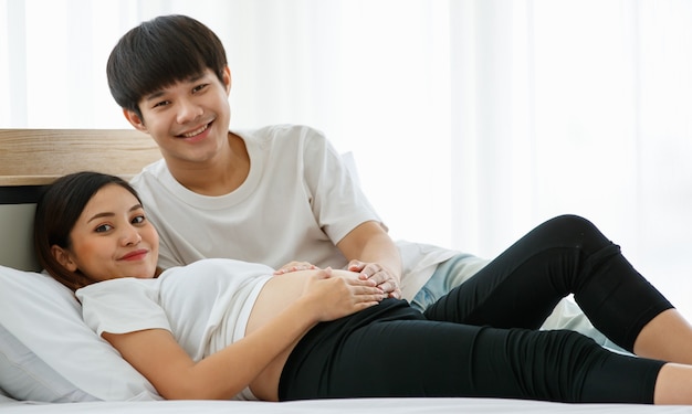 Portrait D'un Beau Jeune Homme Asiatique Et D'une Femme Allongée Sur Un Lit Ensemble. Ils Sourient Joyeusement Et Touchent Le Ventre D'une Mère Enceinte.