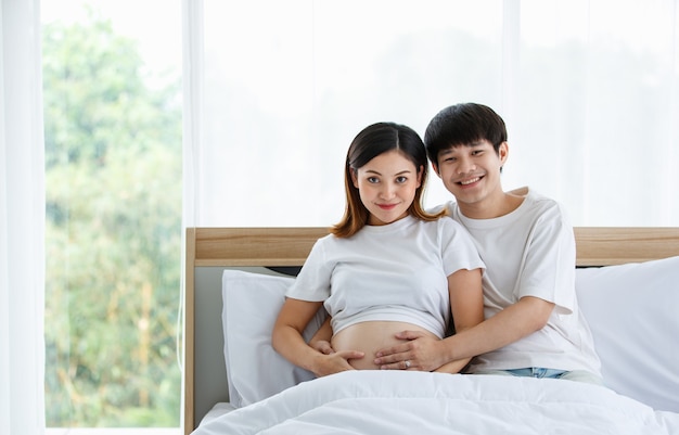 Portrait d'un beau jeune homme asiatique et d'une femme vêtue d'une chemise de nuit blanche assise sur un lit ensemble. Ils sourient joyeusement et touchent le ventre d'une mère enceinte.