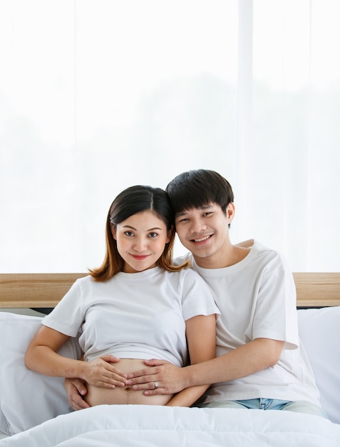 Portrait d'un beau jeune homme asiatique et d'une femme vêtue d'une chemise de nuit blanche assise sur un lit ensemble. Ils sourient joyeusement et touchent le ventre d'une mère enceinte.