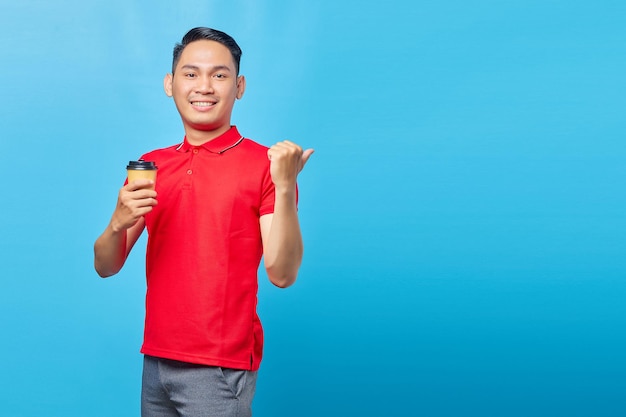 Portrait d'un beau jeune homme asiatique souriant tenant une tasse de café et pointant du doigt l'espace de copie isolé sur fond bleu