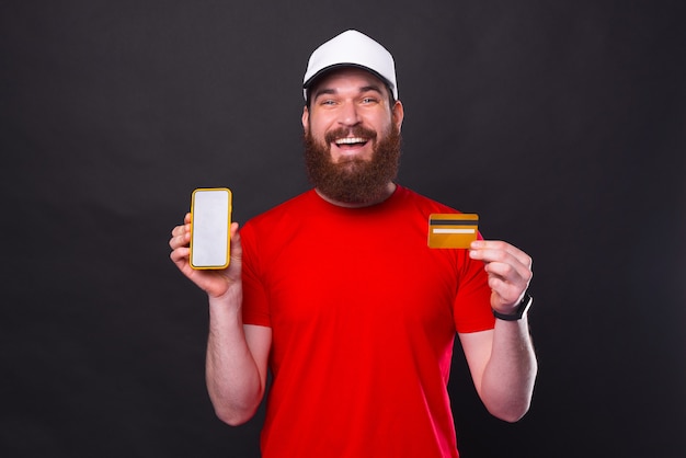 Portrait de beau jeune homme avec barbe montrant smartphone et carte de crédit