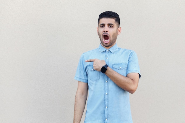 Portrait d'un beau jeune homme barbu choqué en chemise bleue debout pointant sur son côté droit et regardant la caméra avec un visage surpris. studio intérieur tourné isolé sur fond de mur beige clair.