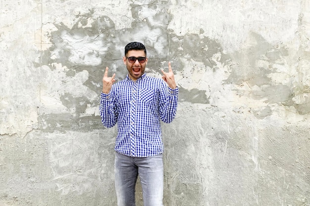 Portrait d'un beau jeune homme barbu fuuny surpris en chemise bleue à carreaux et lunettes de soleil debout contre un mur gris en béton dans un geste de cornes de rock and roll, regardant et criant à la caméra.