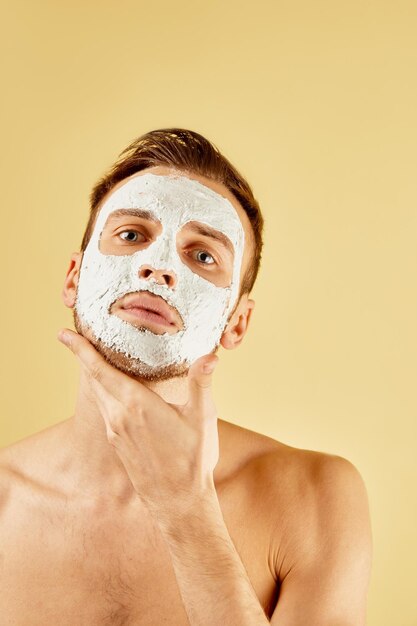 Portrait d'un beau jeune homme barbu avec un masque blanc touche son visage dans le studio isolé sur fond jaune concept de soins de la peau masculine