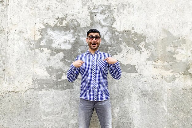 Portrait d'un beau jeune homme barbu surpris en chemise bleue à carreaux et lunettes de soleil debout contre un mur gris béton, se pointant, demandant et regardant avec un visage étonné choqué.