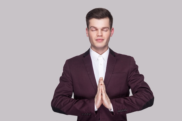 Portrait d'un beau jeune homme calme en costume violet et chemise blanche, debout, yeux fermés avec geste de yoga et méditation. tourné en studio intérieur, isolé sur fond gris.