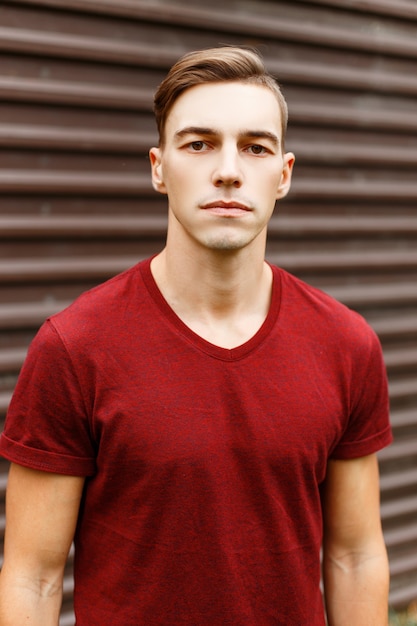 Portrait d'un beau jeune homme cool dans un t-shirt rouge à la mode avec une coiffure élégante près d'un bâtiment en métal vintage