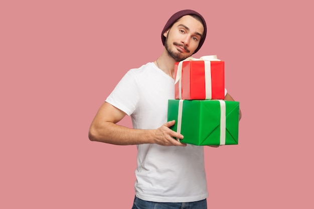 Portrait de beau jeune homme hipster barbu en chemise blanche et chapeau décontracté debout et tenant deux boîte présente, regardant la caméra. Intérieur, isolé, tourné en studio, fond rose