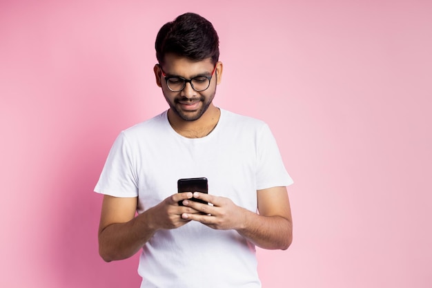 Portrait d'un beau jeune homme indien heureux dans des verres, tenant un téléphone portable, envoyant un message texte, lisant de bonnes nouvelles, posant sur fond rose. Technologie, communication.