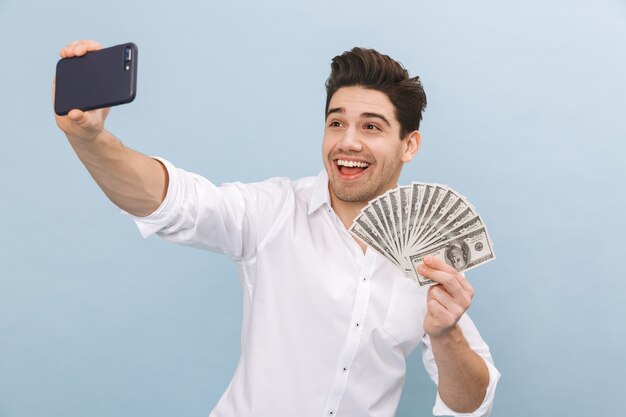 Portrait d'un beau jeune homme joyeux debout isolé sur bleu, montrant des billets d'argent, prenant un selfie