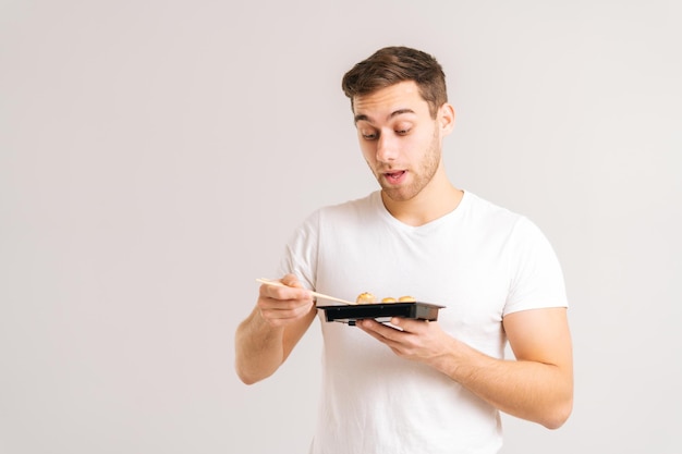 Portrait d'un beau jeune homme mangeant des rouleaux de sushi frais et savoureux avec des baguettes sur fond blanc isolé. Prise de vue en studio d'un homme caucasien heureux qui mange de la nourriture japonaise traditionnelle.