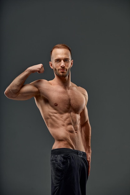 Portrait d'un beau jeune homme modèle avec une coiffure dans un débardeur blanc à la mode maquette avec un corps musclé en bonne santé exhibant ses muscles biceps sur un fond gris