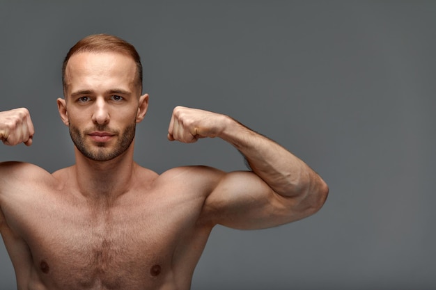 Photo portrait d'un beau jeune homme modèle un torse nu avec un corps sain musclé exhibant ses muscles biceps sur un fond gris