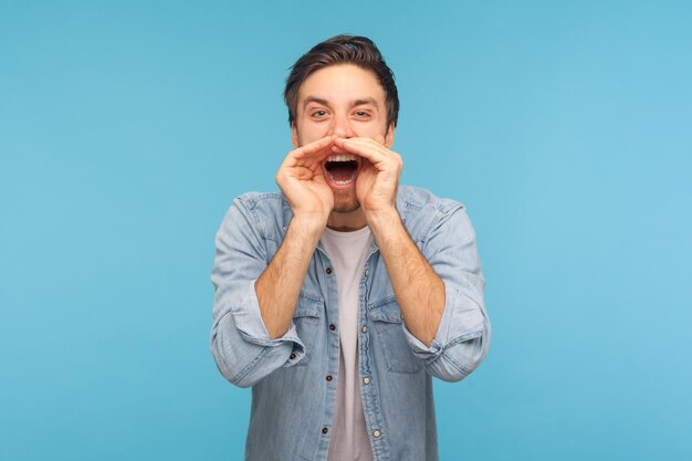 Portrait d'un beau jeune homme portant une chemise en jean criant largement ouvrant la bouche tenant les mains sur le visage criant annonçant son opinion Prise de vue en studio intérieur isolée sur fond bleu