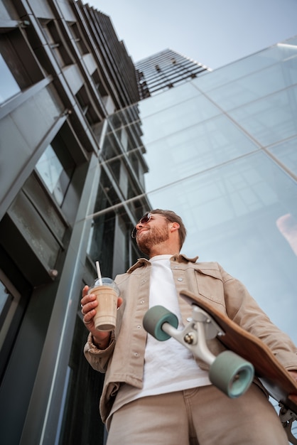 Portrait d'un beau jeune homme portant des lunettes de soleil vue de dessous tenant une tasse de café cappuccino à emporter debout sur un bâtiment en verre de la rue urbaine skateur hipster tenant un longboard.