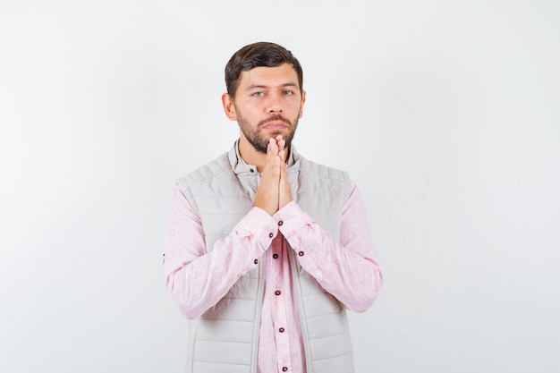 Portrait de beau jeune homme pressant les mains ensemble tout en priant en chemise, gilet et regardant la vue de face silencieuse