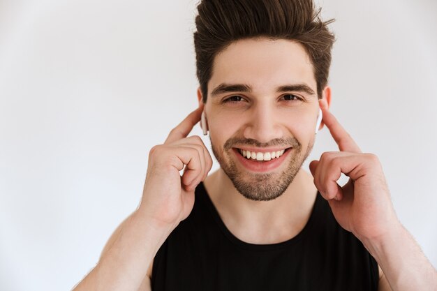 Portrait d'un beau jeune homme sportif souriant et joyeux isolé sur un mur blanc, écoutant de la musique avec des écouteurs.