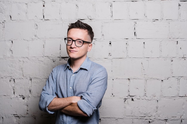 Portrait de beau jeune homme en vêtements jeans et lunettes, debout contre le mur de briques grises