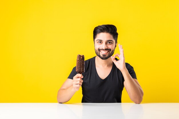 Portrait d'un beau jeune indien barbu mangeant de la crème glacée dans un cône ou un popsicle assis à table sur fond de studio jaune